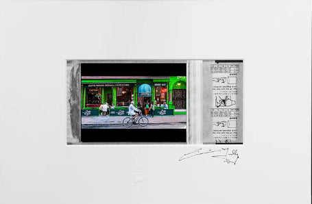 A framed art print featuring a colorful street scene with a cyclist in front of a green storefront, alongside a vertical panel with sketch-like drawings and handwritten notes, all set within a wide white mat and black frame against a dark gray background.