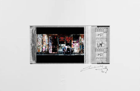 A framed art print or matted photograph displayed against a dark gray background. The print features a large white mat with a central black-and-white photograph showing a colorful storefront scene, alongside a vertical panel with sketches or drawings, plus a signature near the bottom.